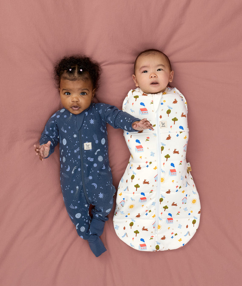 Two babies lying on a pink surface, one wearing a blue slim-fit Long Sleeve Layer, while the other wearing a white Cocoon Swaddle Bag with colourful print.