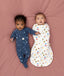 Two babies lying on a pink surface, one wearing a blue slim-fit Long Sleeve Layer, while the other wearing a white Cocoon Swaddle Bag with colourful print.