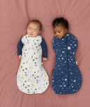 Two babies lying on a pink surface wearing ergoPouch Cocoon Swaddle Bags in 2.5 TOG, one wearing a white Swaddle Bag with both arms out, while the other wearing a blue Swaddle Bag with both arms inside.