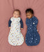 Two babies lying on a pink surface wearing ergoPouch Cocoon Swaddle Bags in 2.5 TOG, one wearing a white Swaddle Bag with both arms out, while the other wearing a blue Swaddle Bag with both arms inside.