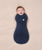 A smiling baby lying on a beige blanket, dressed in a navy blue Cocoon Swaddle Bag 2.5 TOG by ergoPouch, featuring a bell-shaped bottom for natural movement.