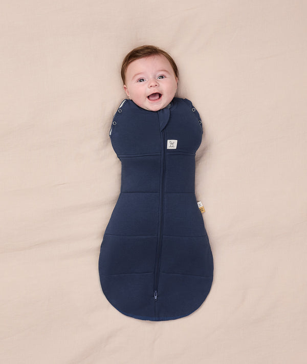 A smiling baby lying on a beige blanket, dressed in a navy blue Cocoon Swaddle Bag 2.5 TOG by ergoPouch, featuring a bell-shaped bottom for natural movement.