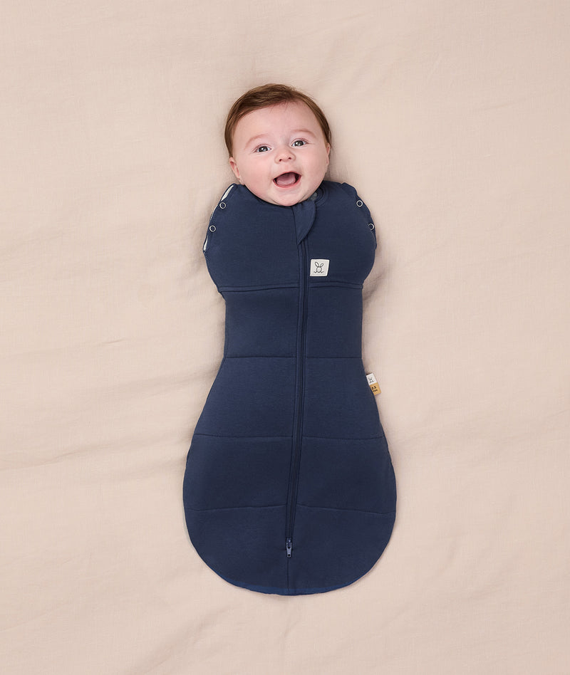 A smiling baby lying on a beige blanket, dressed in a navy blue Cocoon Swaddle Bag 2.5 TOG by ergoPouch, featuring a bell-shaped bottom for natural movement.
