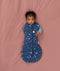 Baby lying on a soft pink surface, wrapped in a blue Cocoon Swaddle Bag with a moon-and-star print, one arm out and looking directly at the camera.