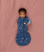 Baby lying on a soft pink surface, wrapped in a blue Cocoon Swaddle Bag with a moon-and-star print, one arm out and looking directly at the camera.