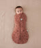 Baby is snugly wrapped in a Cocoon Swaddle Bag, a zip-up swaddle bag in a warm, earthy color. The fabric features a delicate woodland print with small white flowers and dark animal silhouettes like deer and rabbits. The swaddle has a central vertical zipper and a signature bell-shaped bottom to allow for healthy hip movement.
