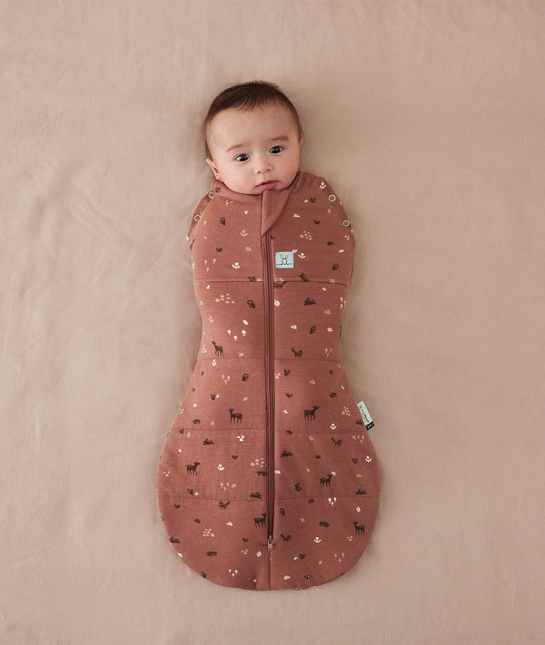 Baby is snugly wrapped in a Cocoon Swaddle Bag, a zip-up swaddle bag in a warm, earthy color. The fabric features a delicate woodland print with small white flowers and dark animal silhouettes like deer and rabbits. The swaddle has a central vertical zipper and a signature bell-shaped bottom to allow for healthy hip movement.