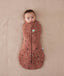 Baby is snugly wrapped in a Cocoon Swaddle Bag, a zip-up swaddle bag in a warm, earthy color. The fabric features a delicate woodland print with small white flowers and dark animal silhouettes like deer and rabbits. The swaddle has a central vertical zipper and a signature bell-shaped bottom to allow for healthy hip movement.