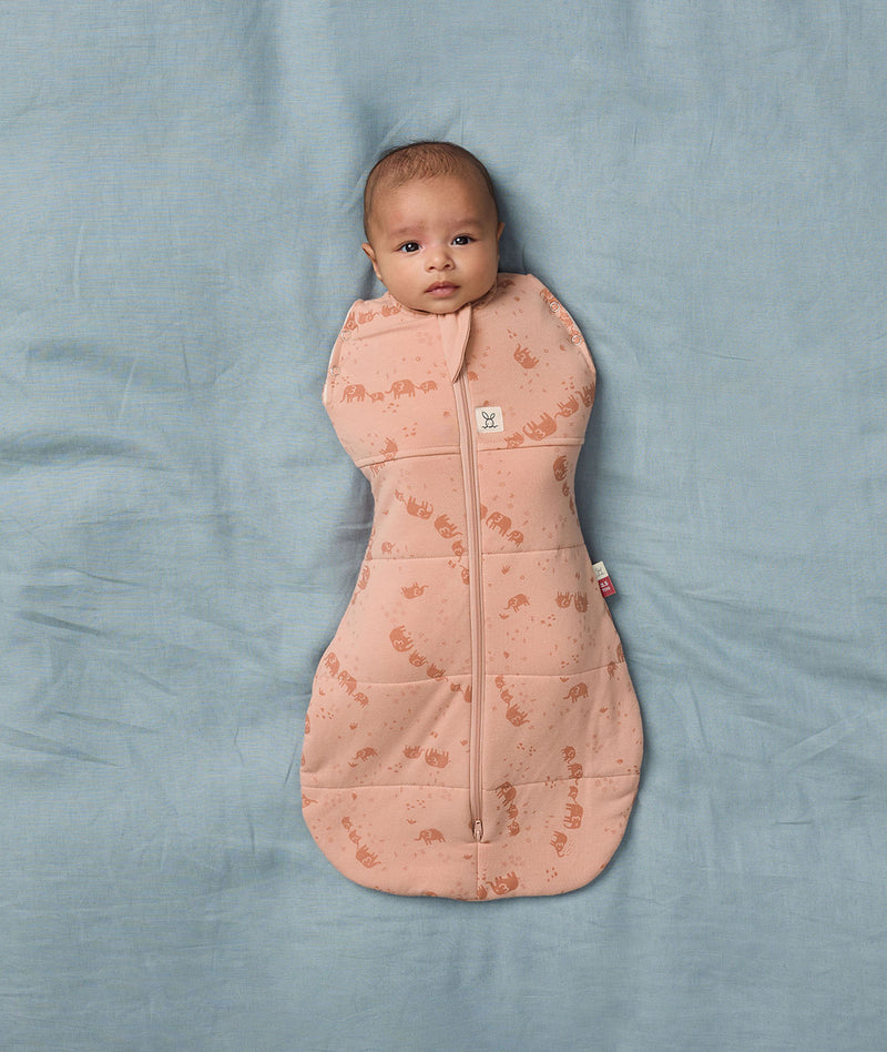 Baby lying on a soft blue sheet wearing an ergoPouch Cocoon Swaddle Bag in Elephant Parade with its arms securely swaddle inside.