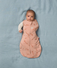 A baby lies on a soft blue sheet, dressed in an ergoPouch Cocoon Swaddle Bag with its one arm outside.