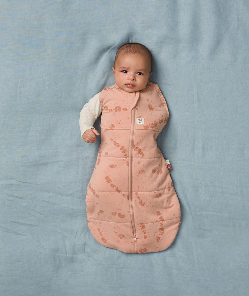A baby lies on a soft blue sheet, dressed in an ergoPouch Cocoon Swaddle Bag with its one arm outside.