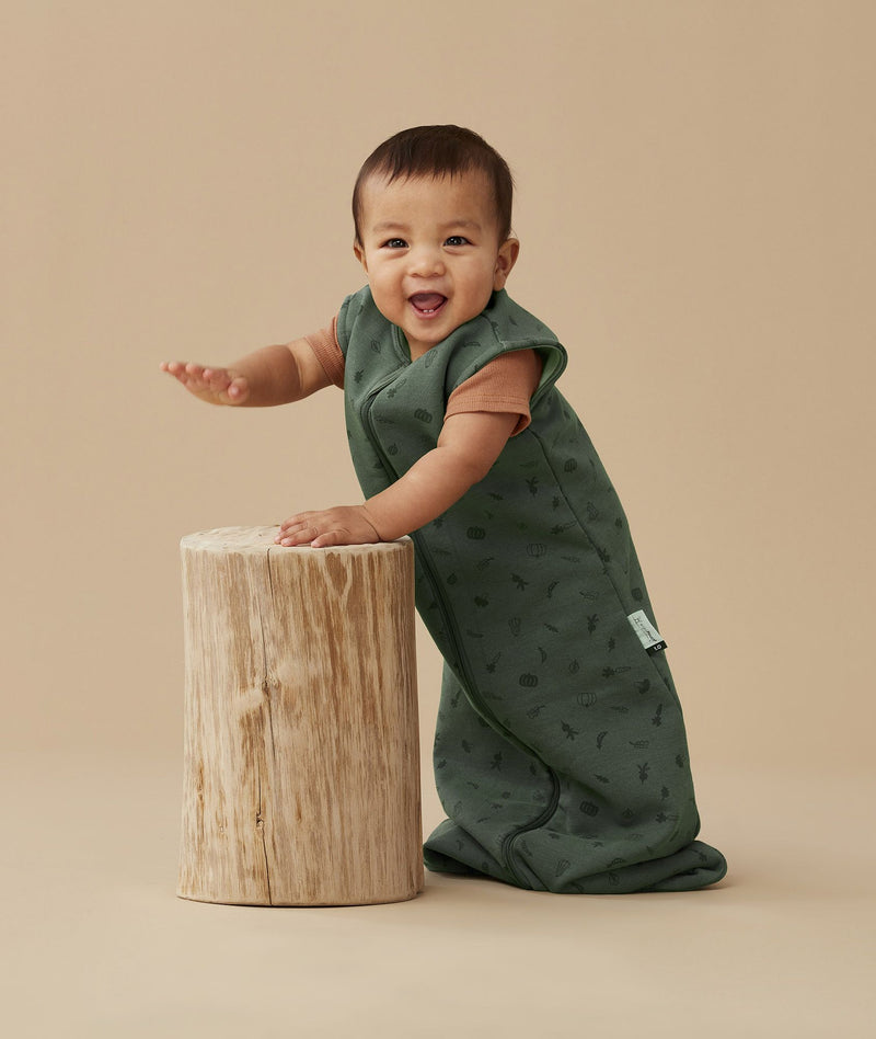 Baby standing and smiling while leaning on a wooden stump, wearing a green sleeping bag with veggie patterns.