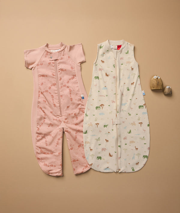 Two baby sleep suits laid flat on a beige surface. The left suit is pink with short sleeves and an elephant print, featuring a zipper down the front and separated legs. The right suit is sleeveless, cream-colored with a multicolored animal print, also with a front zipper.
