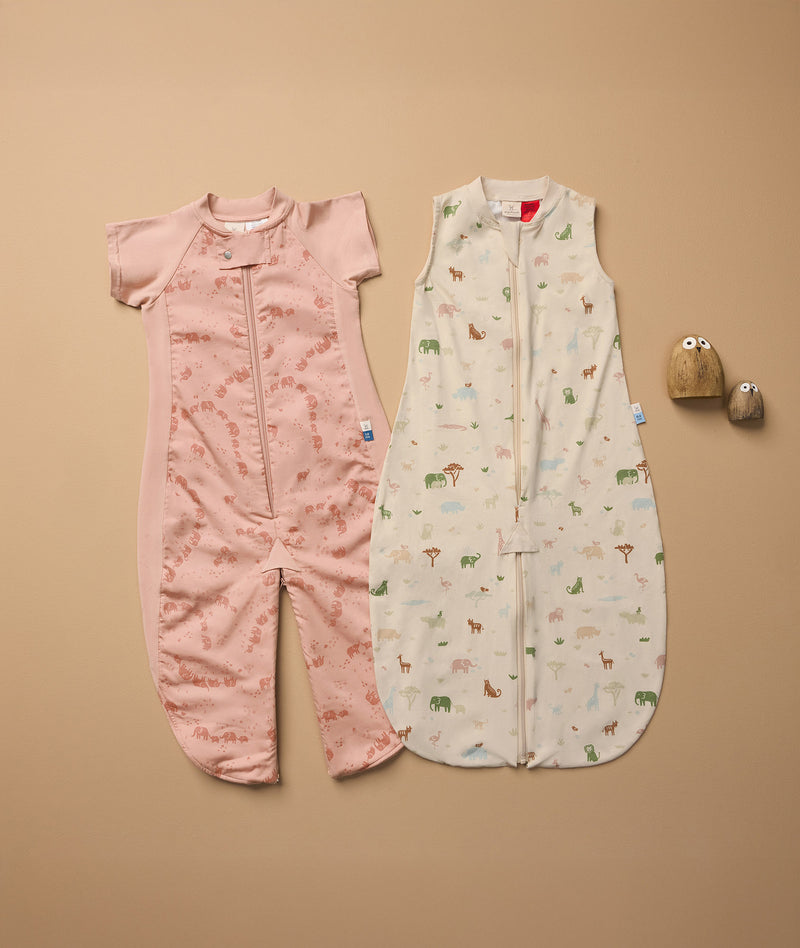 Two baby sleep suits laid flat on a beige surface. The left suit is pink with short sleeves and an elephant print, featuring a zipper down the front and separated legs. The right suit is sleeveless, cream-colored with a multicolored animal print, also with a front zipper.