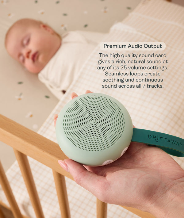 An adult hand holds a round, mint-green Drift Away white noise machine next to a cot where a baby is peacefully sleeping. A text box highlights 