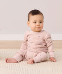 An infant sits on a rug wearing ergoPouch Long Sleeve Baby Pyjamas in Daisies, which feature shoulder press studs for easy wear.