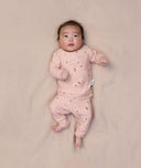 A baby is lying on a light pink sheet, wearing ergoPouch two-piece long sleeve baby pyjamas in Daisies.
