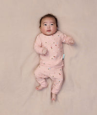 A baby is lying on a light pink sheet, wearing ergoPouch two-piece long sleeve baby pyjamas in Daisies.