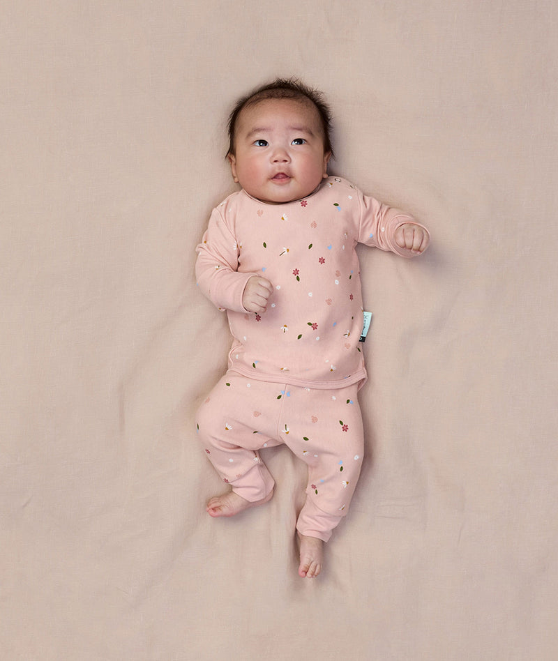 A baby is lying on a light pink sheet, wearing ergoPouch two-piece long sleeve baby pyjamas in Daisies.