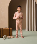 A preschooler stands next to a vintage suitcase and wooden toys, wearing peach ergoPouch long sleeve pyjamas featuring a snug fit and a cute elephant print.