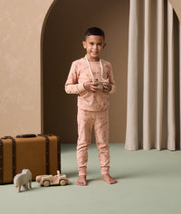 A preschooler stands next to a vintage suitcase and wooden toys, wearing peach ergoPouch long sleeve pyjamas featuring a snug fit and a cute elephant print.