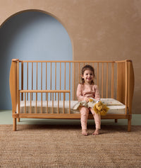 A toddler is sitting on a wooden crib with an open side, smiling and holding a plush lion toy. The child is wearing pink long-sleeve pyjamas with a subtle animal print pattern. The pajamas consist of a fitted top and matching bottoms, both made from soft, comfortable fabric.