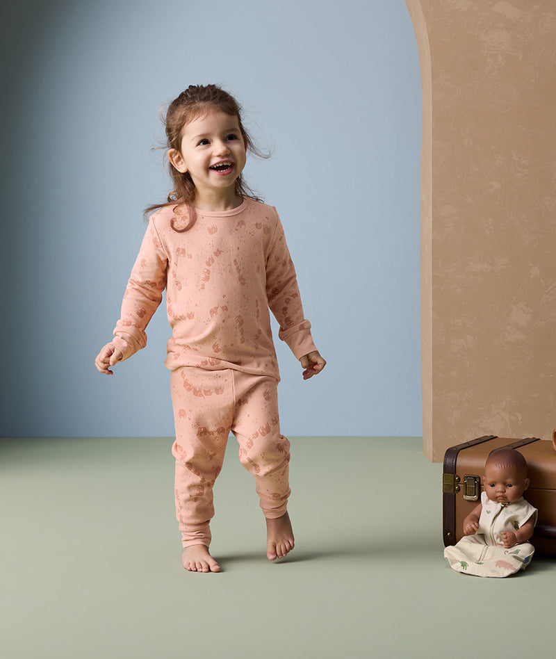 A cheerful toddler is wearing peach-colored long sleeve pyjamas from ergoPouch, featuring an elephant print and a snug fit.