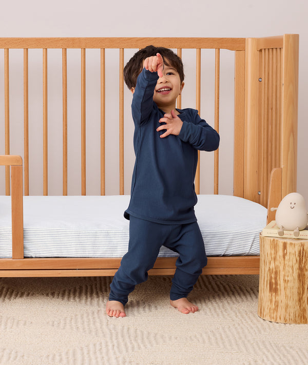Toddler standing and playing beside a toddler bed, wearing a navy blue long-sleeve pyjama set with matching pants, featuring a relaxed fit and soft fabric.
