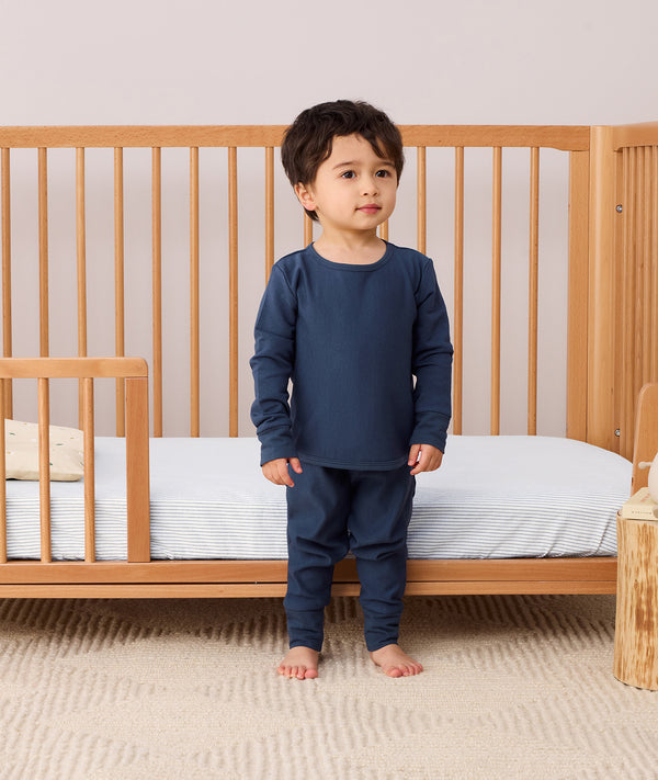 Toddler standing in front a crib wearing a navy blue long-sleeve pyjama set with matching pants, made from soft organic cotton and bamboo.