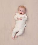 A baby lying on a soft beige surface, wearing a long-sleeve, cream-colored pyjama set with a snug fit and a shoulder snap.