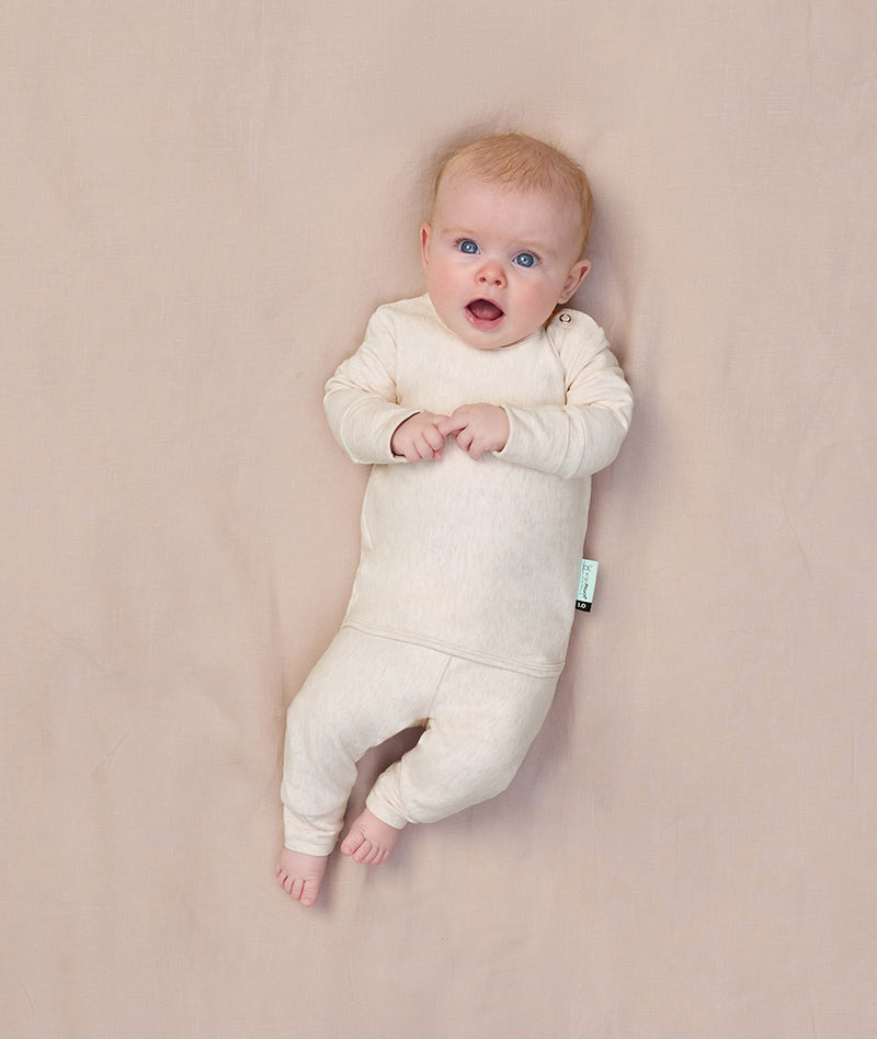 A baby lying on a soft beige surface, wearing a long-sleeve, cream-colored pyjama set with a snug fit and a shoulder snap.