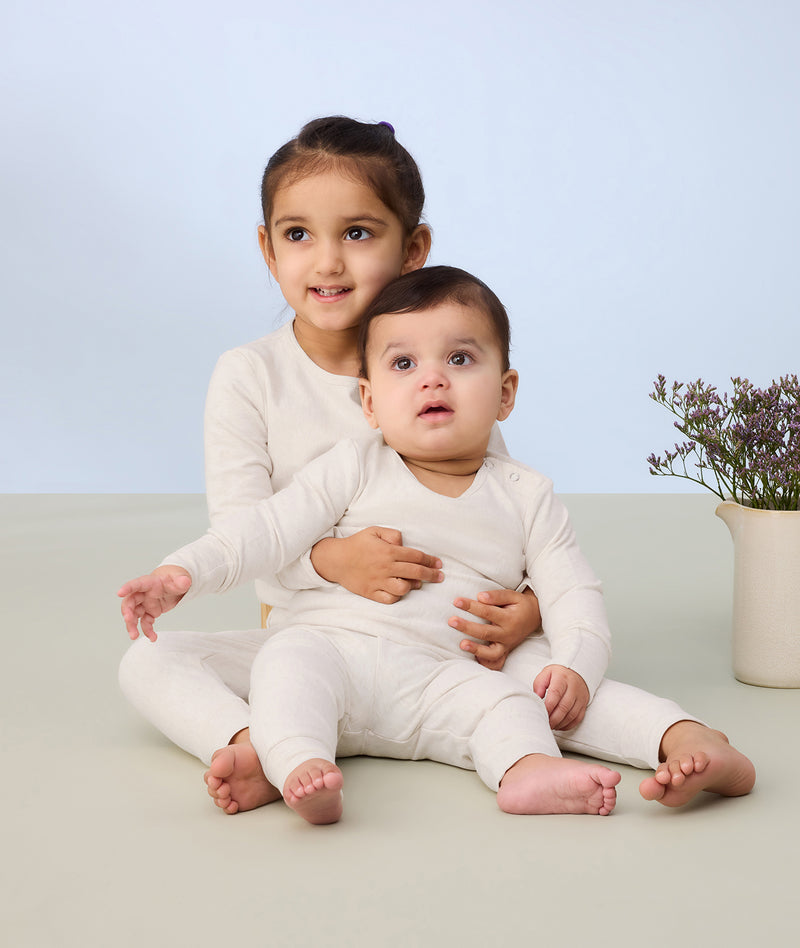  A young child and a baby sitting together on a light-colored surface, both wearing matching long-sleeve, cream-colored pyjamas. The pyjamas have a soft, snug fit with covered legs and minimal detailing