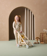 A preschooler is joyfully pushing a wooden toy stroller with a plush lion inside. She is wearing cream-colored long-sleeve pyjamas with safari animal print pattern.