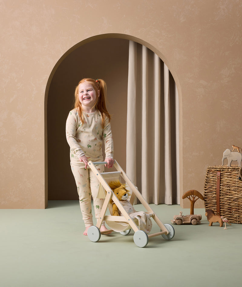 A preschooler is joyfully pushing a wooden toy stroller with a plush lion inside. She is wearing cream-colored long-sleeve pyjamas with safari animal print pattern.
