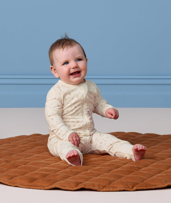 Smiling baby sitting on a brown mat, wearing a cream patterned Long Sleeve Layer 0.2 TOG