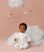 Baby wearing a white slim-fit Long Sleeve Layer sitting upright against a light pink background with decorative birds and clouds.