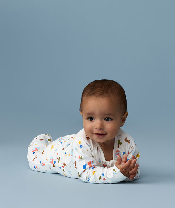 Infant lying on its stomach against a blue background wearing a white Long Sleeve Layer with colourful all-over print.