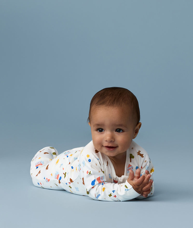 Infant lying on its stomach against a blue background wearing a white Long Sleeve Layer with colourful all-over print.