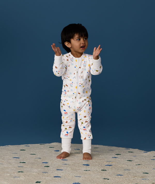 Toddler standing on a rug against a deep blue background wearing a white Long Sleeve Layer, featuring a front zipper and colourful all-over print.