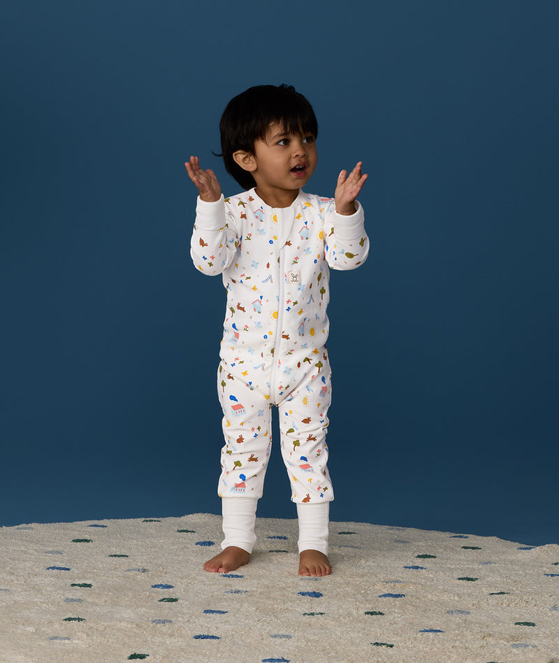 Toddler standing on a rug against a deep blue background wearing a white Long Sleeve Layer, featuring a front zipper and colourful all-over print.