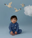 Baby sitting on a light blue background wearing a blue Long Sleeve Layer with a moon-and-star print, looking toward the camera, with decorative birds and a cloud above.