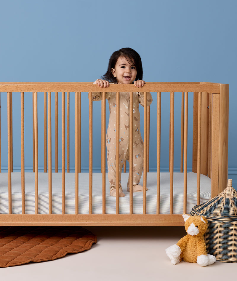 Toddler standing up in a wooden cot wearing a patterned tan Long Sleeve  Layer.