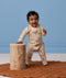 Baby wearing a patterned tan Long-Sleeve Layer leaning against a wooden stool.