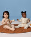 Two infants sitting on a brown mat, wearing ergoPouch Long Sleeve Layer in Melody and Lullaby.