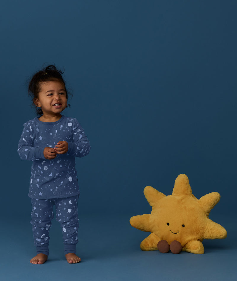 Toddler wearing a blue Long Sleeve Pyjama with star and moon print standing barefoot on a blue background with a sun plush toy on the side