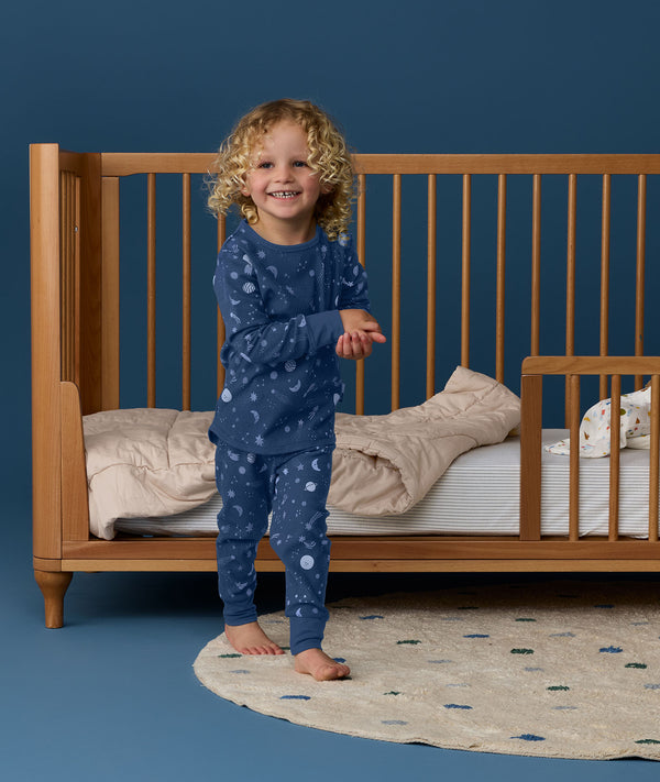 Young child wearing blue Long Sleeve Pyjamas with a moon-and-star print, standing barefoot beside a wooden cot and smiling at the camera.