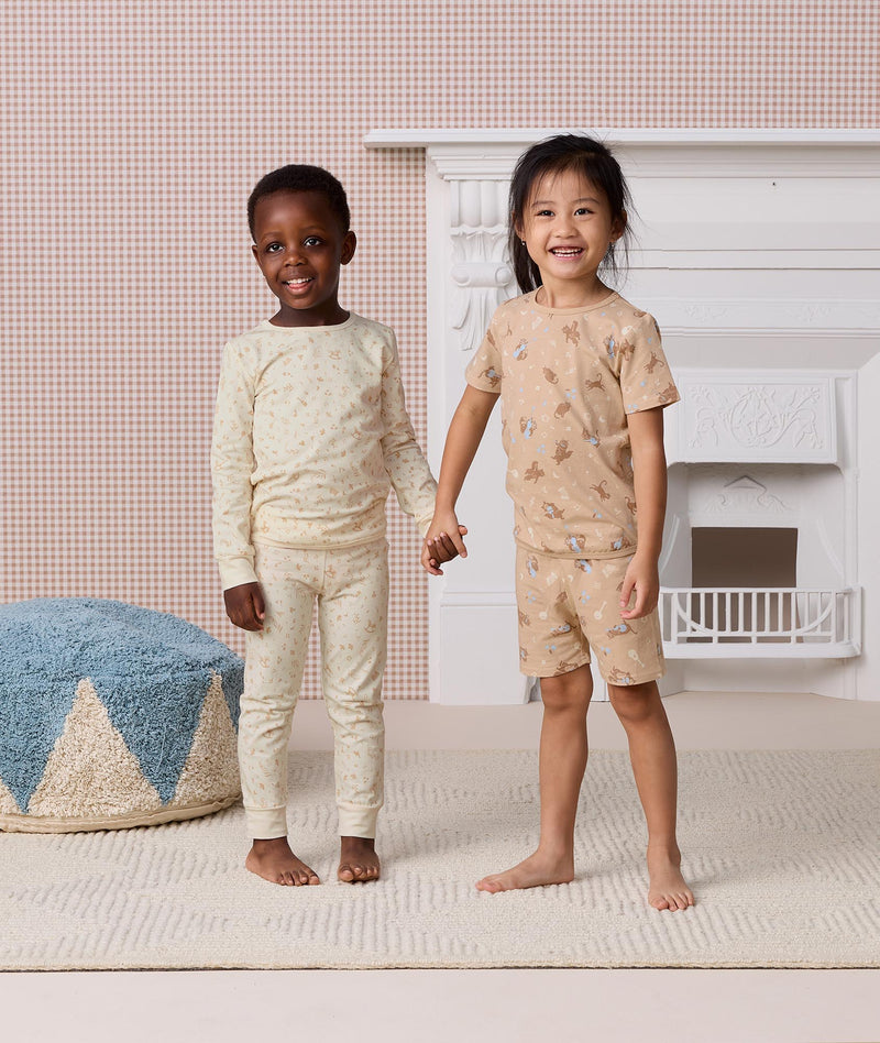 Two preschoolers holding hands, wearing ergoPouch Pyjama Sets. The young boy is wearing a cream Long Sleeve Pyjama Set, while the girl is wearing a tan Short Sleeve Pyjama Set.