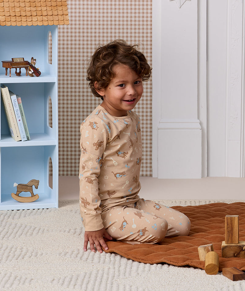 Smiling child sitting on the floor in patterned beige Long Sleeve Pyjamas next to a dollhouse.
