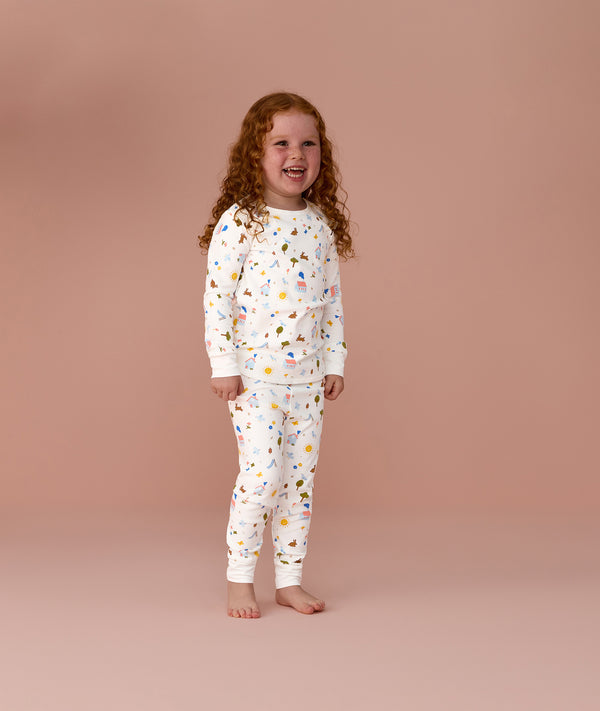 Preschooler wearing a white long sleeve pyjamas with colourful all-over print, standing barefoot against a pink background.