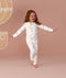 Young child wearing white long-sleeve pyjamas with a colorful all-over print, standing barefoot against a soft pink background with arms outstretched.